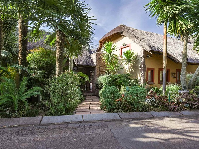 A charming front entrance featuring a traditional thatched roof, stone-clad accents, and a paved walkway surrounded by lush, established garden landscaping.