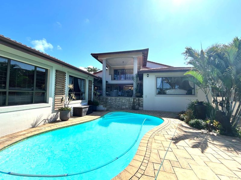 A well-maintained outdoor pool area featuring a paved deck, stone-clad accents, and a covered patio with an upper-level balcony.