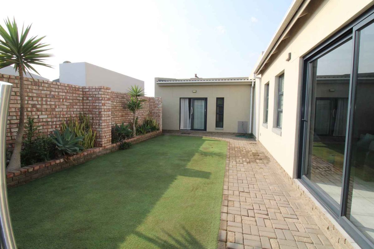 A well-maintained rear garden featuring a paved walkway, artificial lawn, and a brick boundary wall, providing access to the home via sliding glass doors.