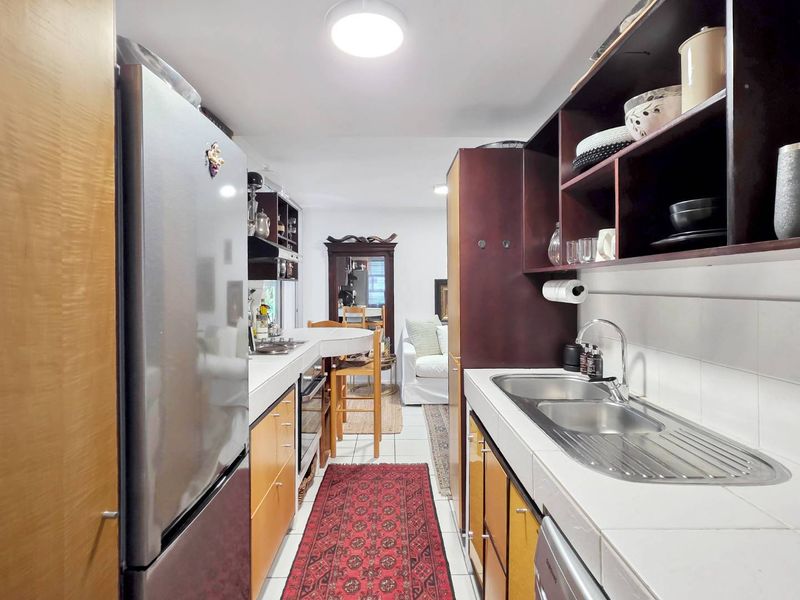 A galley-style kitchen featuring tiled countertops, double stainless steel sinks, and dark wood cabinetry with open shelving, leading into an adjacent living area.