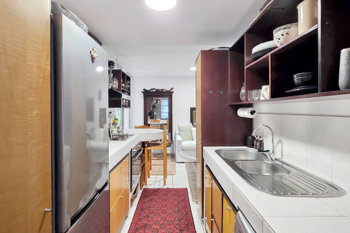 A galley-style kitchen featuring tiled countertops, double stainless steel sinks, and dark wood cabinetry with open shelving, leading into an adjacent living area.