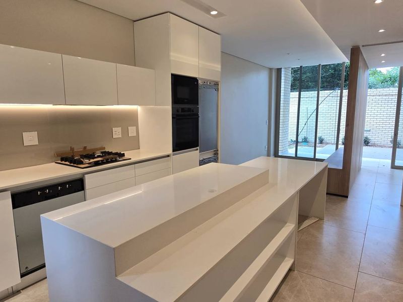 Modern, high-end kitchen featuring sleek white cabinetry, stone countertops, integrated appliances, and a central island, leading to an outdoor patio area.