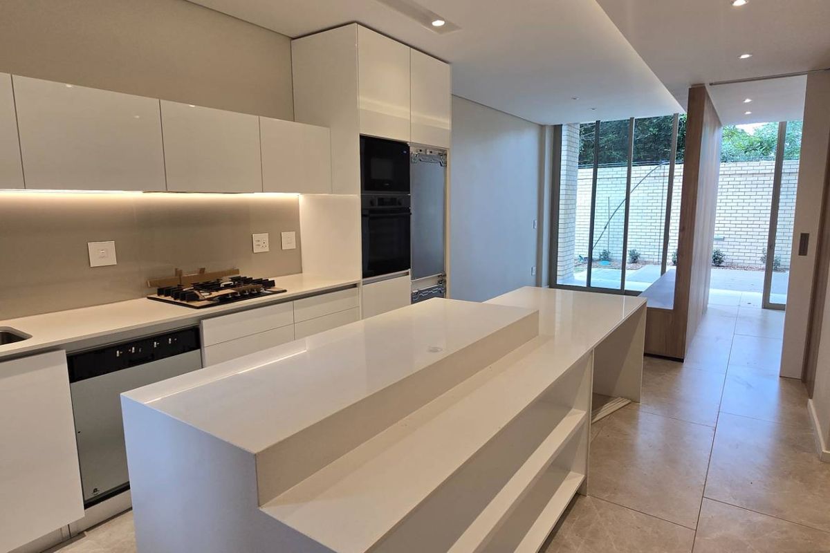 Modern, high-end kitchen featuring sleek white cabinetry, stone countertops, integrated appliances, and a central island, leading to an outdoor patio area.