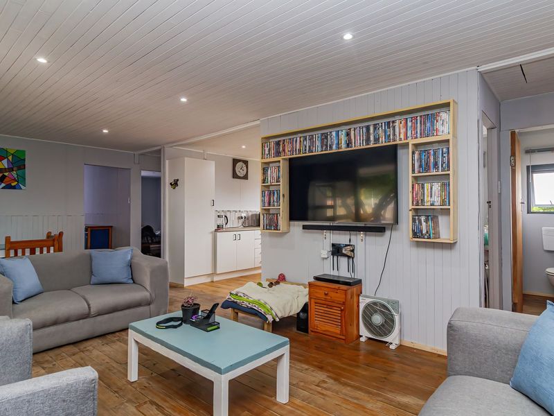 Open plan living area featuring timber flooring and tongue and groove ceiling, with an integrated kitchen and built-in shelving unit.