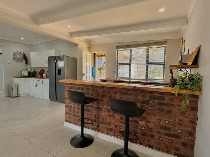 Open plan kitchen and living area featuring a rustic exposed brick breakfast bar, white cabinetry, and neutral tiled flooring throughout.