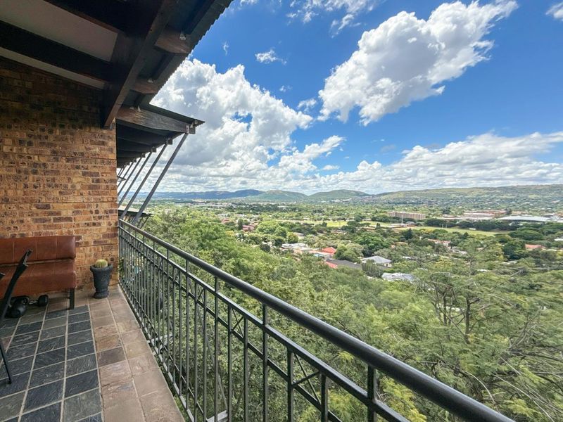 A covered balcony featuring exposed brickwork, tiled flooring, and metal railings, offering an expansive elevated view of the surrounding landscape.