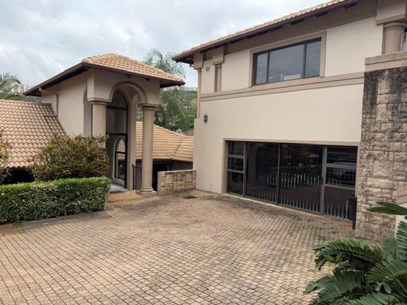 Front exterior view of the townhouse featuring a paved driveway, stone cladding accents, arched entrance with columns, and a multi-level tiled roof design.