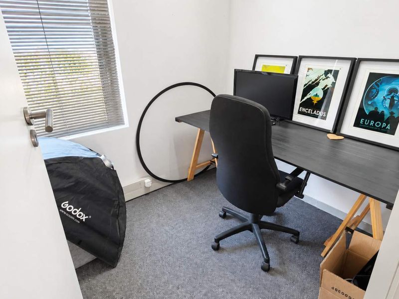 A compact office space featuring grey carpet flooring, a window with venetian blinds, and standard interior door hardware.