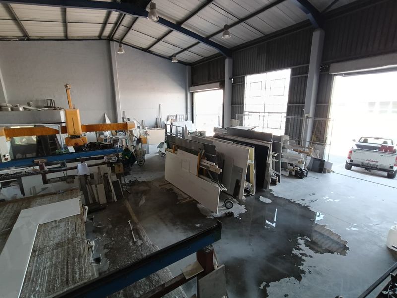 Industrial warehouse space featuring high ceilings, concrete flooring, and large roller shutter access, currently utilized for stone fabrication and material storage.