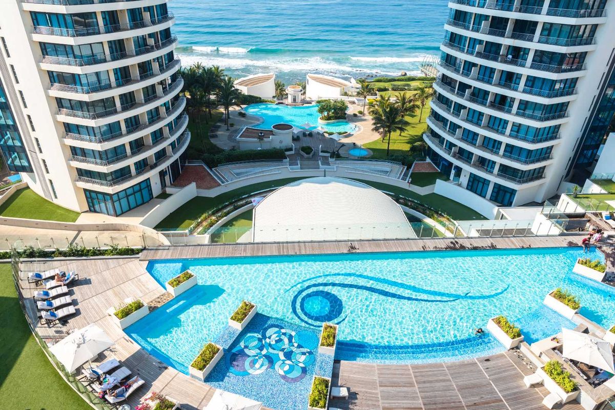 Aerial view of a luxury apartment complex featuring a large swimming pool with decorative tiling, landscaped grounds, and direct ocean views.
