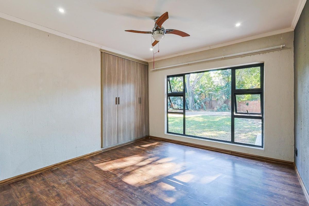 A bright, empty bedroom featuring laminate flooring, built-in cupboards, a ceiling fan, and a large window overlooking the garden.