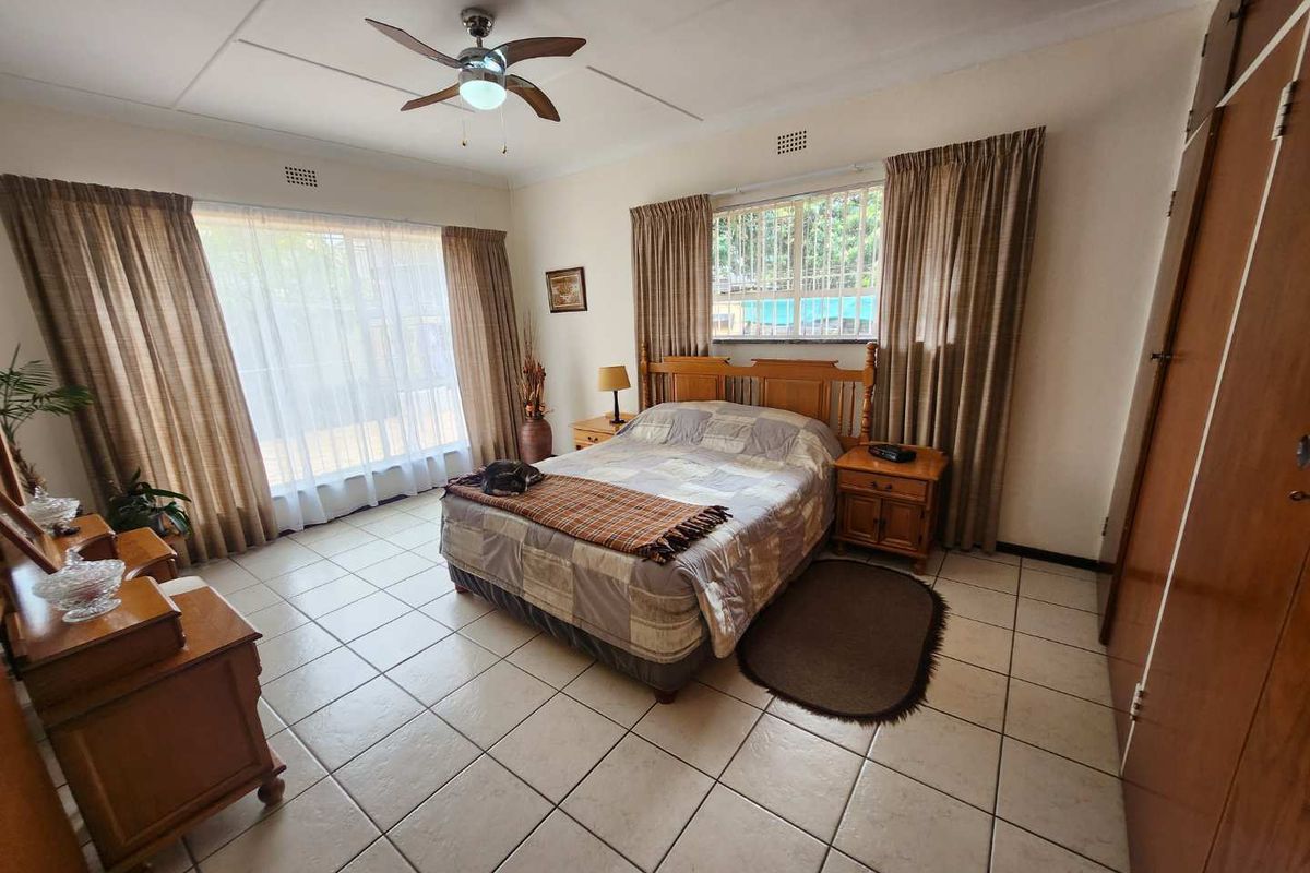 A spacious bedroom featuring tiled flooring, built-in wooden cupboards, a ceiling fan, and large windows with curtains providing natural light.