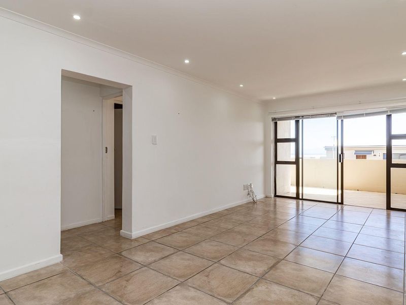 A bright, empty living room featuring neutral tiled flooring, recessed downlighting, and a large sliding glass door providing access to an outdoor balcony.