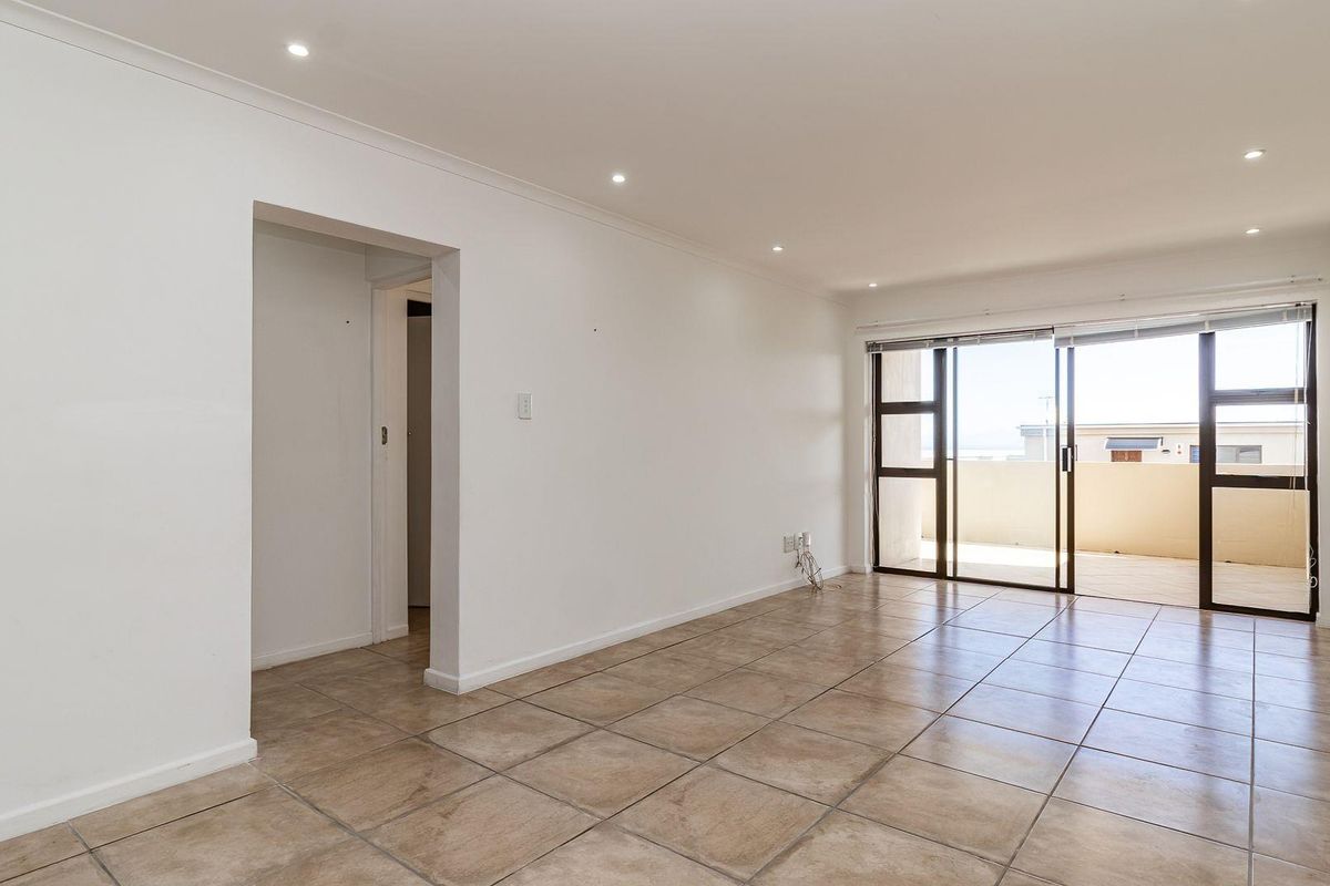 A bright, empty living room featuring neutral tiled flooring, recessed downlighting, and a large sliding glass door providing access to an outdoor balcony.