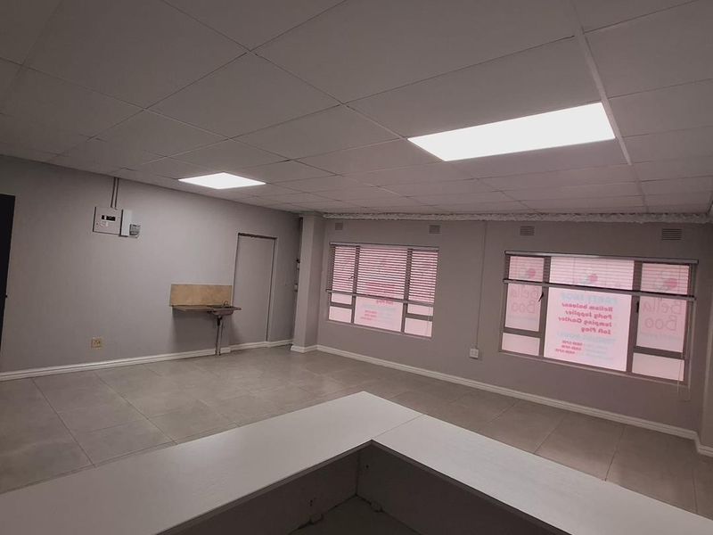 Spacious open-plan office area featuring tiled flooring, a suspended ceiling with recessed lighting, large windows, and a small built-in kitchenette sink.