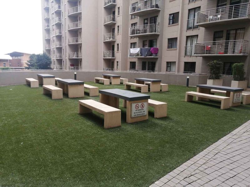 Communal outdoor terrace area featuring artificial grass, fixed concrete seating, and paved walkways, situated within an apartment complex.