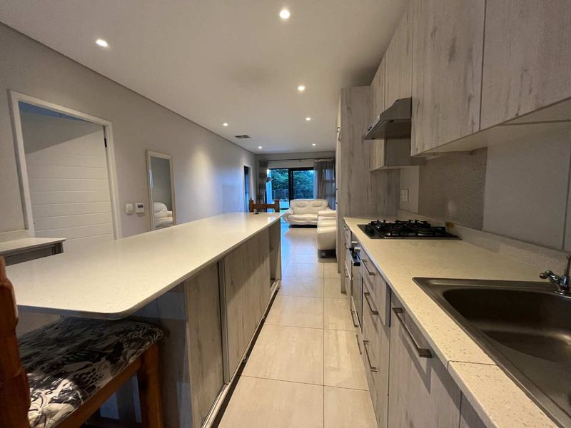 Modern open-plan kitchen featuring light wood-grain cabinetry, stone countertops, gas hob, and integrated extractor fan, flowing into the living area.