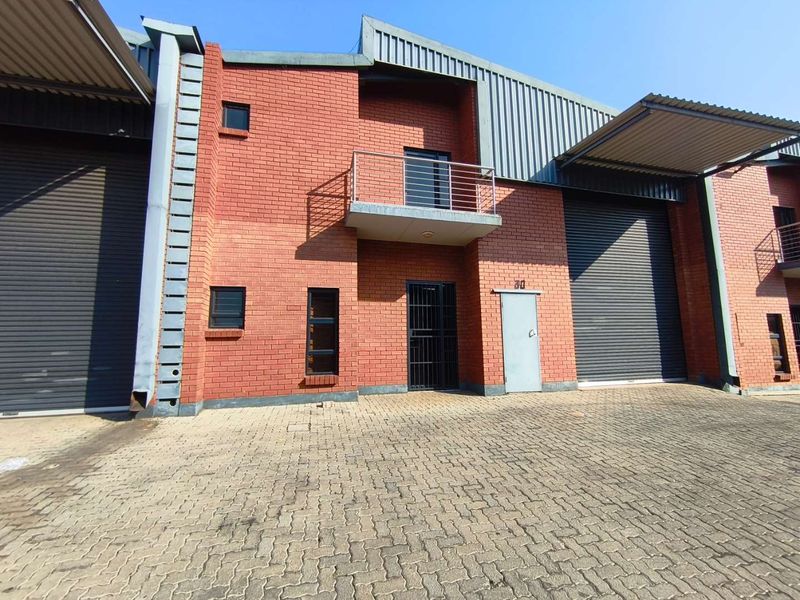 Front elevation of an industrial unit featuring red brick construction, roller shutter doors, a small balcony, and a paved driveway area.