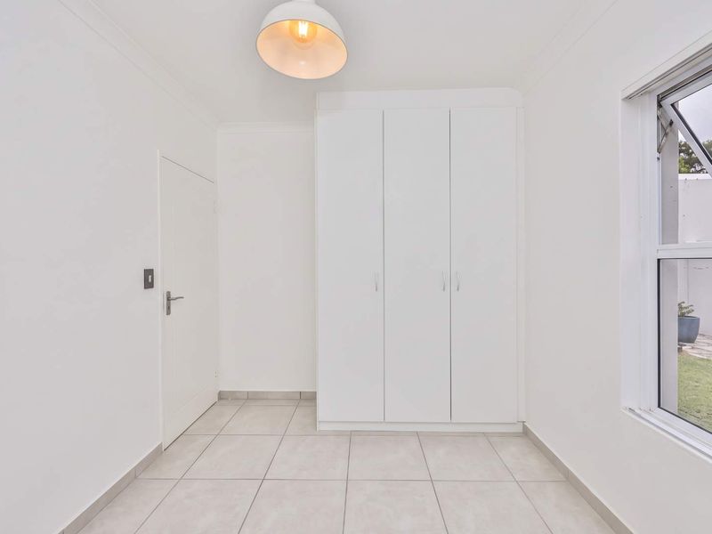 A bright, empty bedroom featuring neutral tiled flooring, a built-in white wardrobe, and a window overlooking the garden.