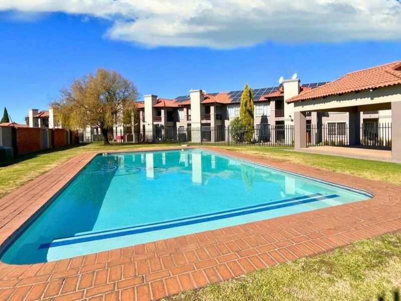 A well-maintained communal swimming pool area featuring brick coping, surrounding lawn, and a covered patio structure within an apartment complex.