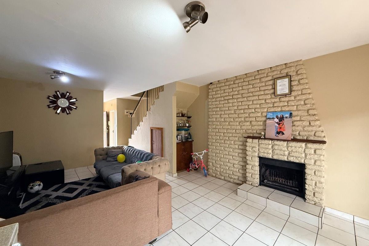Spacious living room featuring a prominent brick fireplace, tiled flooring, and an integrated staircase, offering a functional layout with potential for modernization.