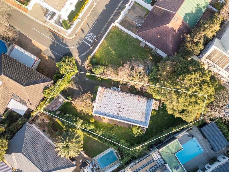 Aerial view of a residential property featuring a detached house with a corrugated metal roof, situated on a corner plot with surrounding garden space.