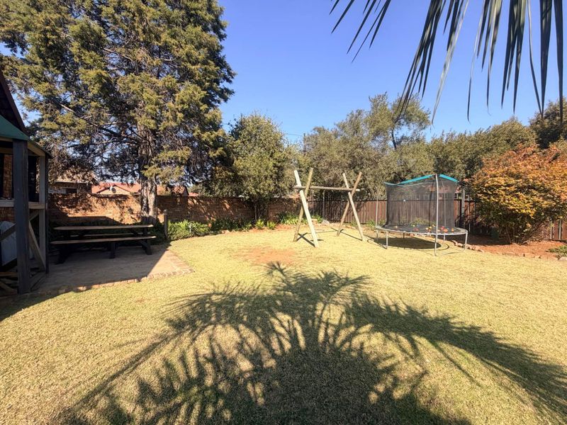 A spacious private backyard featuring a well-maintained lawn, mature trees, a paved patio area, and brick boundary walls.