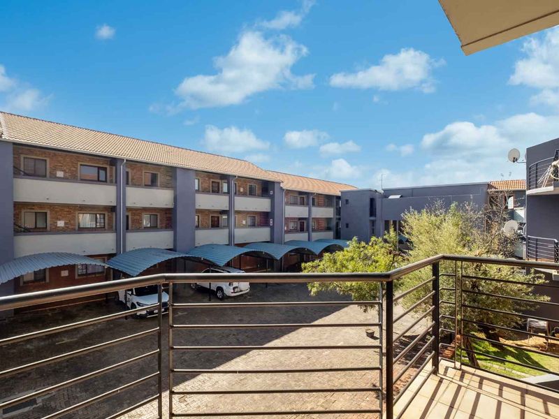 A view from a balcony overlooking the apartment complex courtyard, featuring paved parking areas with covered carports and established trees.