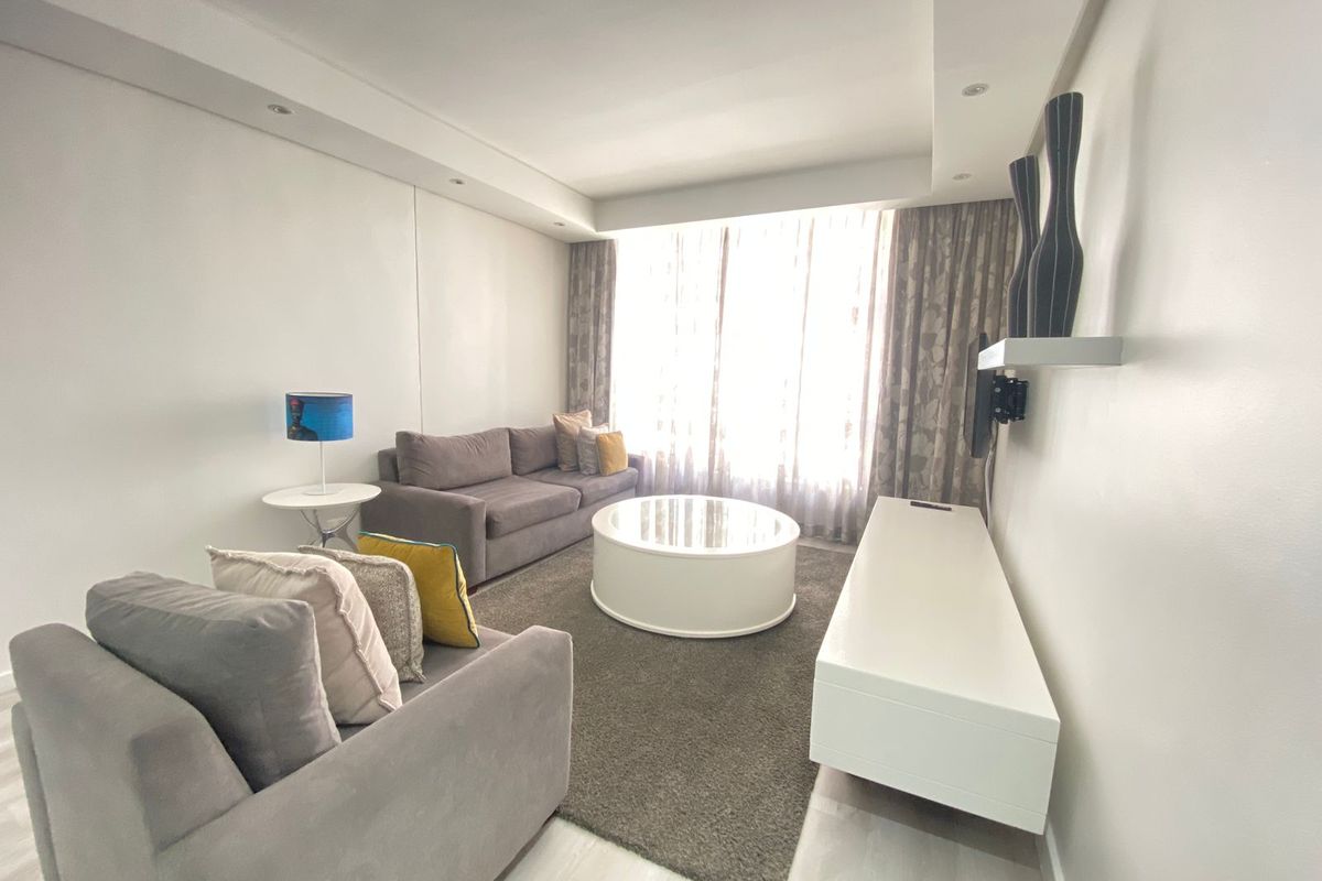 A bright and modern living room featuring recessed ceiling lighting, laminate flooring, and a clean, neutral aesthetic with large windows.