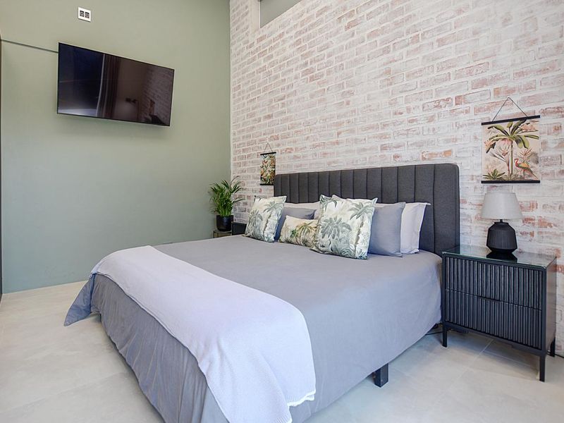 A modern bedroom featuring a textured exposed brick feature wall, neutral tiled flooring, and a wall-mounted television.