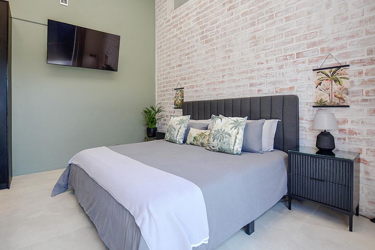A modern bedroom featuring a textured exposed brick feature wall, neutral tiled flooring, and a wall-mounted television.