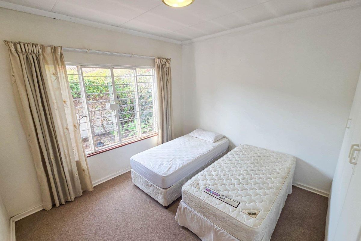 A functional bedroom featuring carpeted flooring, a built-in cupboard, and a large window fitted with security bars.