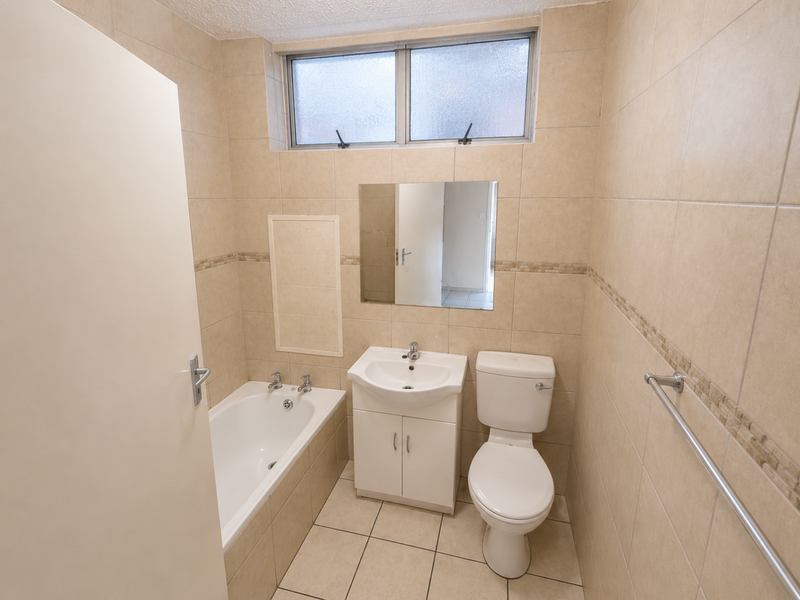 A functional bathroom featuring a bathtub, vanity cabinet with basin, toilet, and neutral wall and floor tiling with a frosted glass window.