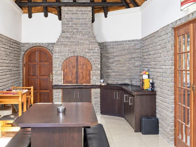 Spacious enclosed patio featuring a traditional thatched roof, exposed timber rafters, built-in brick braai, and integrated cabinetry with a sink.