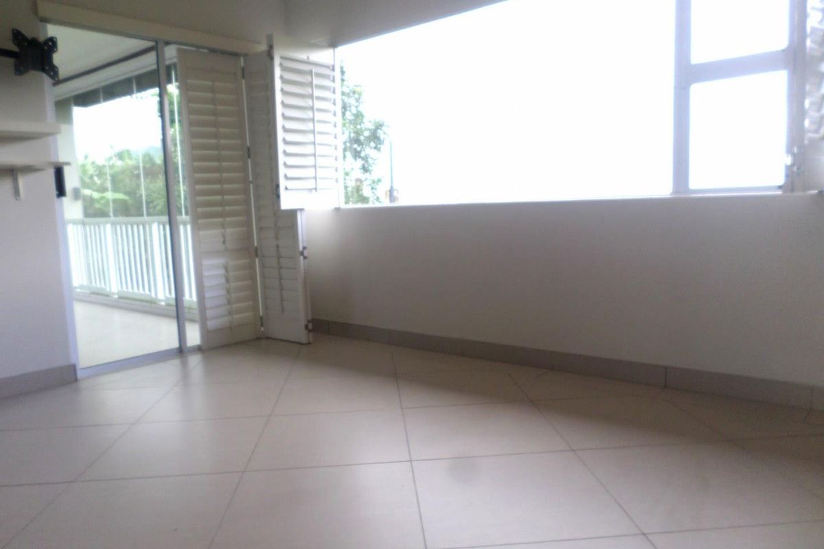 A bright, empty living area featuring large windows, tiled flooring, and plantation shutters, with direct access to an outdoor balcony.