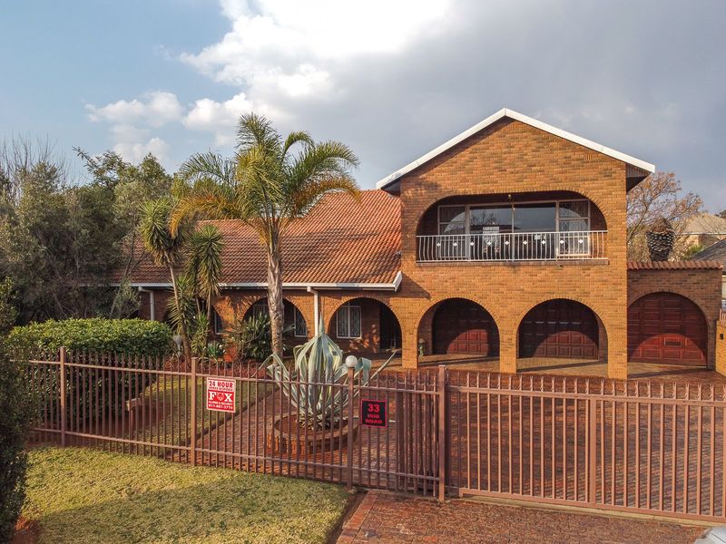 A well-maintained face brick family home featuring arched garage bays, an upper-level balcony, and secure perimeter fencing with a paved driveway.