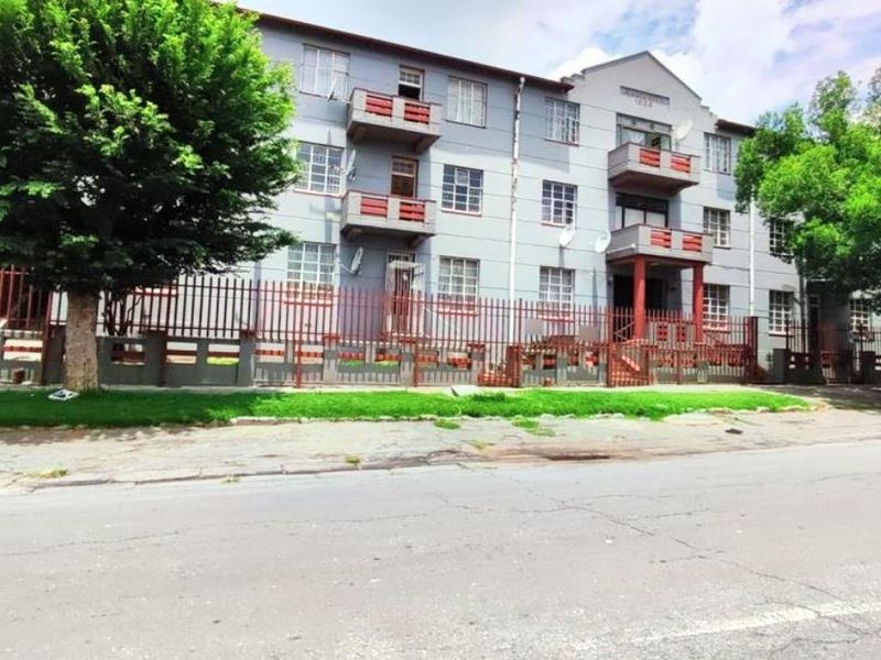 Street view of a multi-story apartment building featuring a metal perimeter fence, balconies, and a paved sidewalk.
