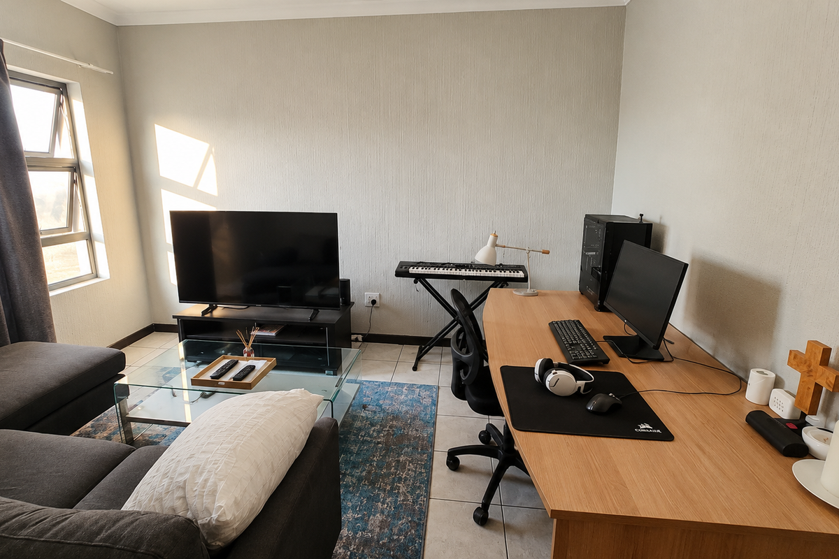 A functional living room featuring tiled flooring and natural light from an aluminium-framed window, currently utilized as a combined lounge and workspace area.