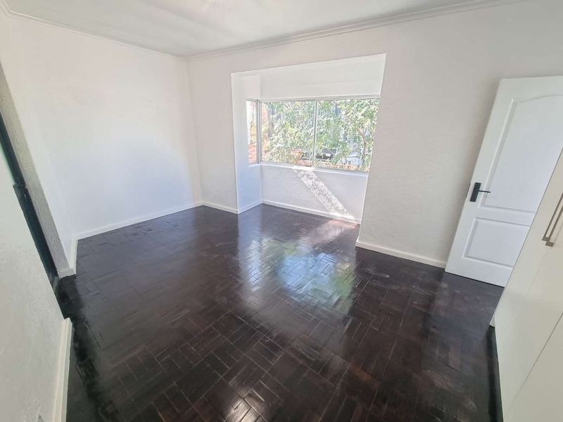 A bright, empty bedroom featuring dark polished parquet flooring, a built-in cupboard, and a large window providing natural light.