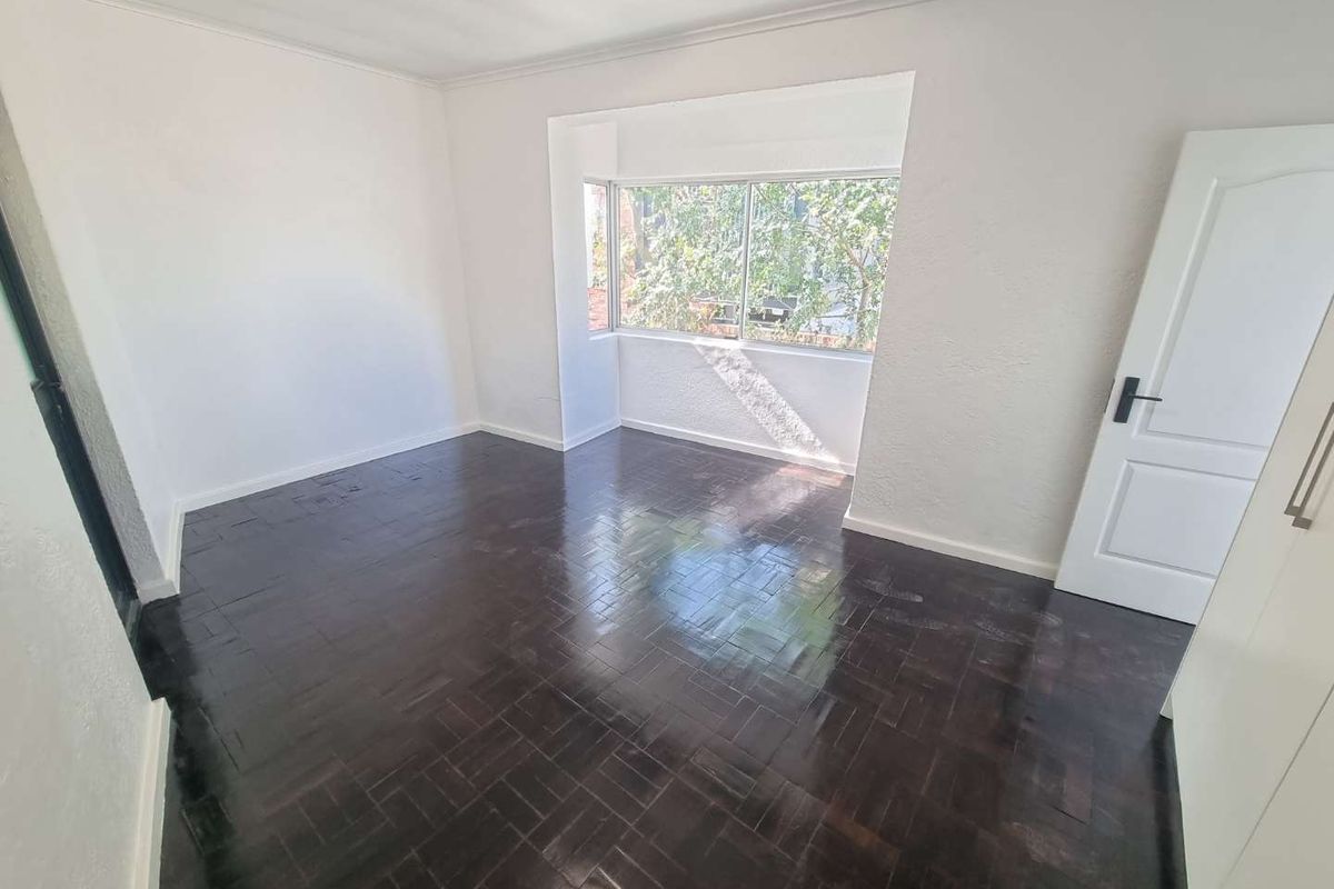 A bright, empty bedroom featuring dark polished parquet flooring, a built-in cupboard, and a large window providing natural light.