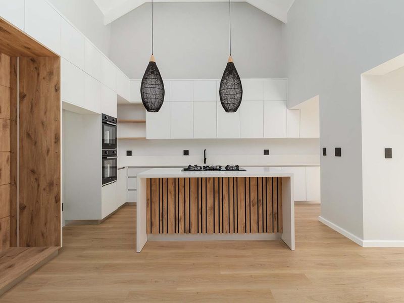 Modern, high-end kitchen featuring a central island with gas hob, built-in ovens, sleek white cabinetry, and a striking vaulted ceiling.