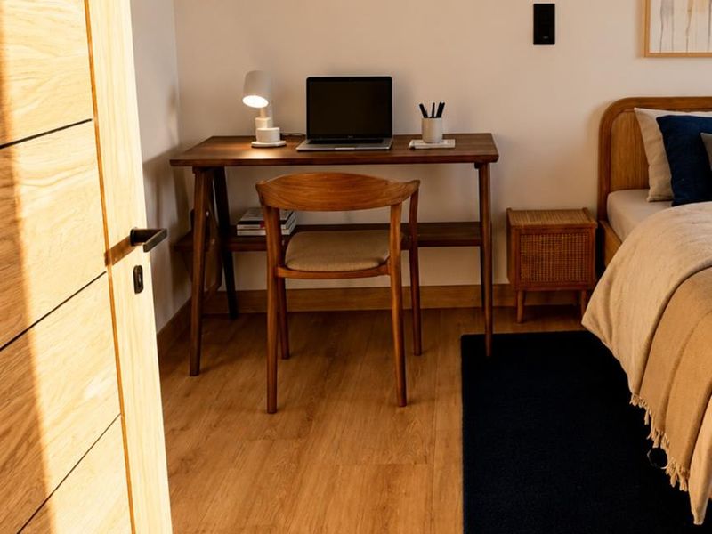 A well-lit bedroom featuring light wood flooring, a modern timber door, and a dedicated workspace area.