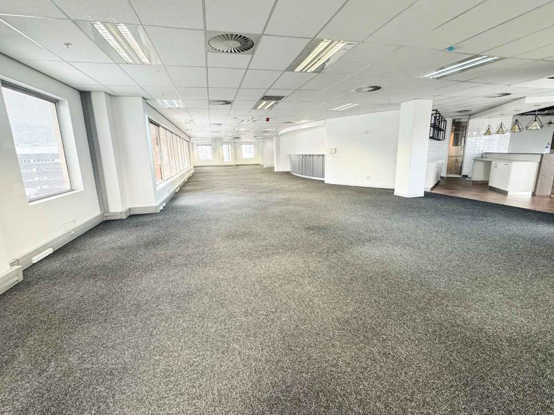 Spacious open-plan office area featuring carpeted flooring, suspended ceilings with recessed lighting, large windows for natural light, and a small integrated kitchenette.