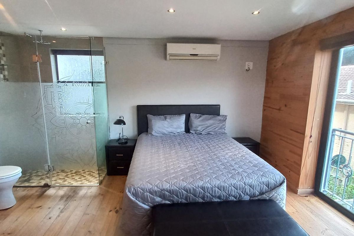 A modern bedroom featuring an open-plan en-suite bathroom with a glass-enclosed shower, timber flooring, and sliding door access to a balcony.