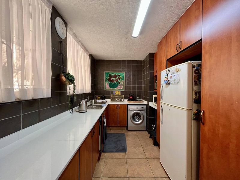 A functional galley-style kitchen featuring dark tiled walls, wooden cabinetry, white countertops, and integrated laundry appliance space.