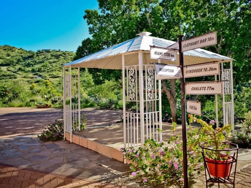 A scenic outdoor garden area featuring a white metal gazebo set on stone paving, surrounded by lush greenery and directional signage.