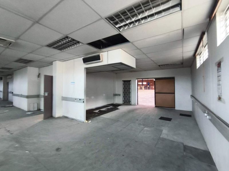 An empty industrial office space featuring a suspended ceiling, recessed lighting, and direct access to a larger warehouse area.