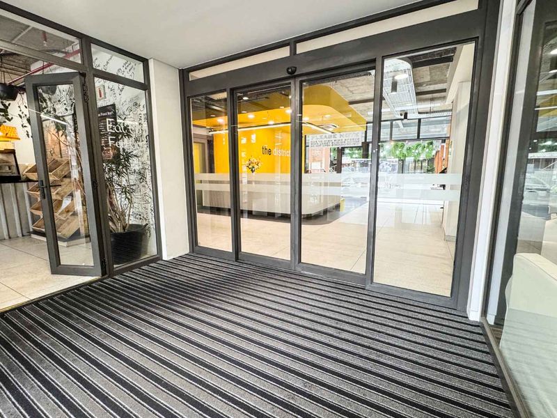 Modern commercial building entrance featuring automatic sliding glass doors, durable recessed matting, and clean tiled flooring with an open-plan interior view.
