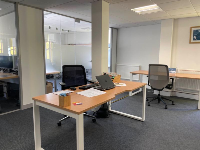 A professional office space featuring glass partition walls, recessed ceiling lighting, and carpet flooring, providing a bright and functional work environment.