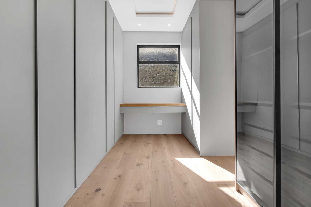 A modern, minimalist walk-in closet or dressing room featuring extensive built-in cabinetry, light timber flooring, and a sleek integrated desk area.